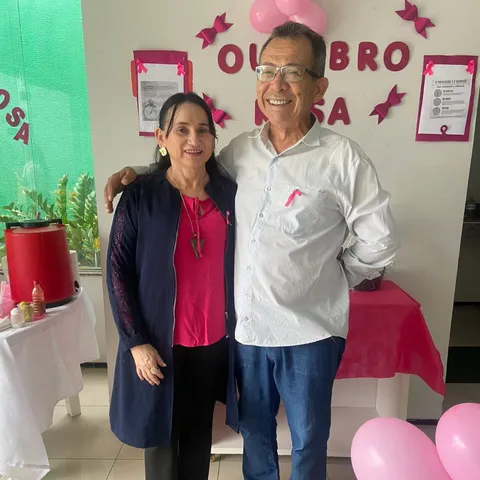 Uma mulher e um homem posam diante da decoração do Outubro Rosa, com letras e balões cor-de-rosa.