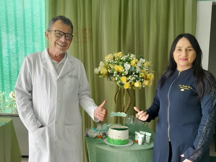 Um homem de jaleco branco e uma mulher de casaco azul-marinho sorriem e fazem sinal de positivo ao lado de uma mesa com bolo e flores.