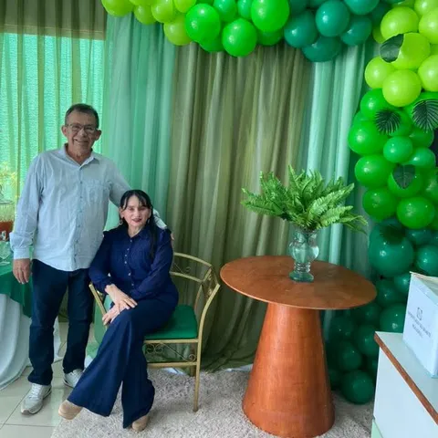 Um homem em pé e uma mulher sentada, sob um arco de balões verdes.