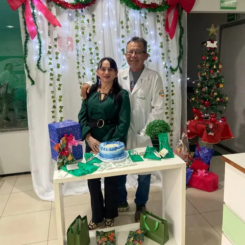 Uma mulher de vestido verde e um homem de jaleco branco atrás de uma mesa com bolo e presentes, numa decoração de Natal.