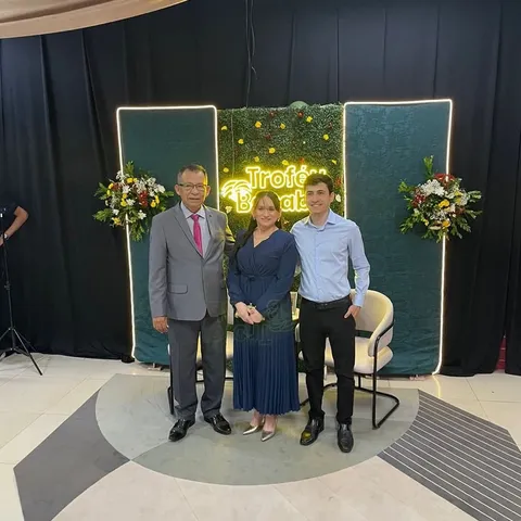 Uma mulher posa com homens diante de um letreiro luminoso, com flores dos lados.